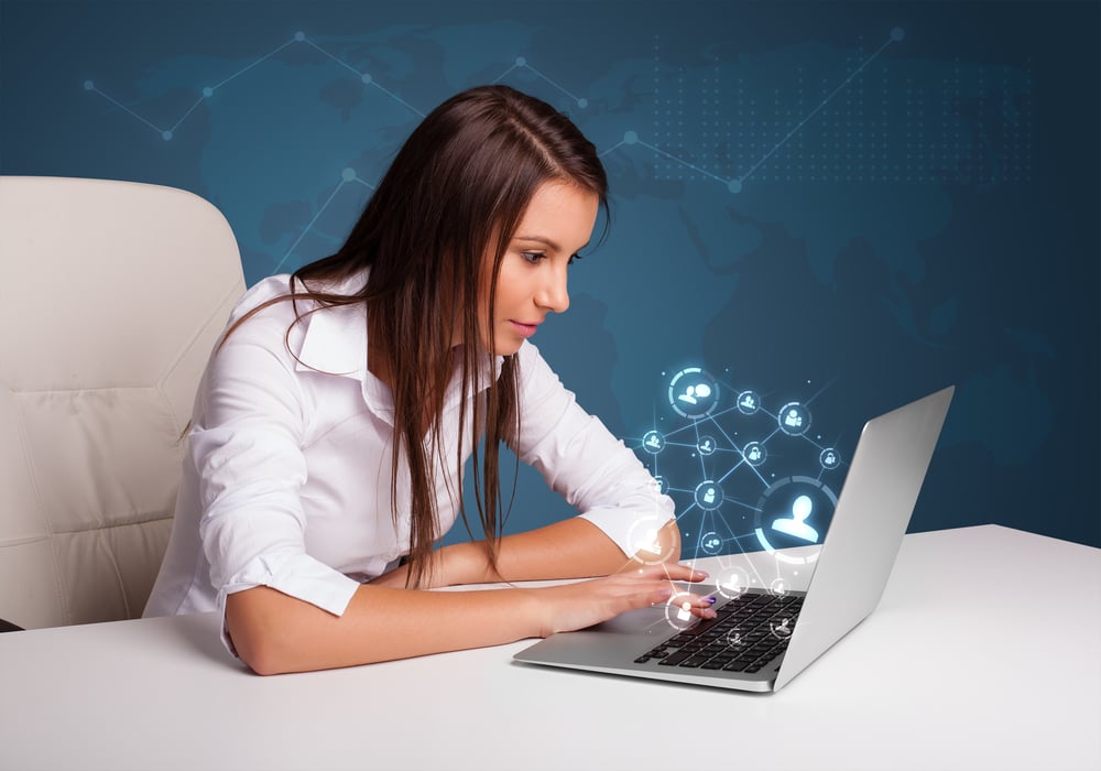 Beautiful young lady sitting at desk and typing on laptop with social network icons comming out