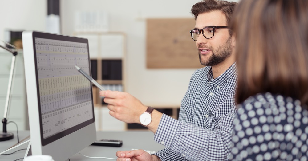 Proficient young male employee with eyeglasses and checkered shirt, explaining a business analysis displayed on the monitor of a desktop PC to his female colleague, in the interior of a modern office-1-1